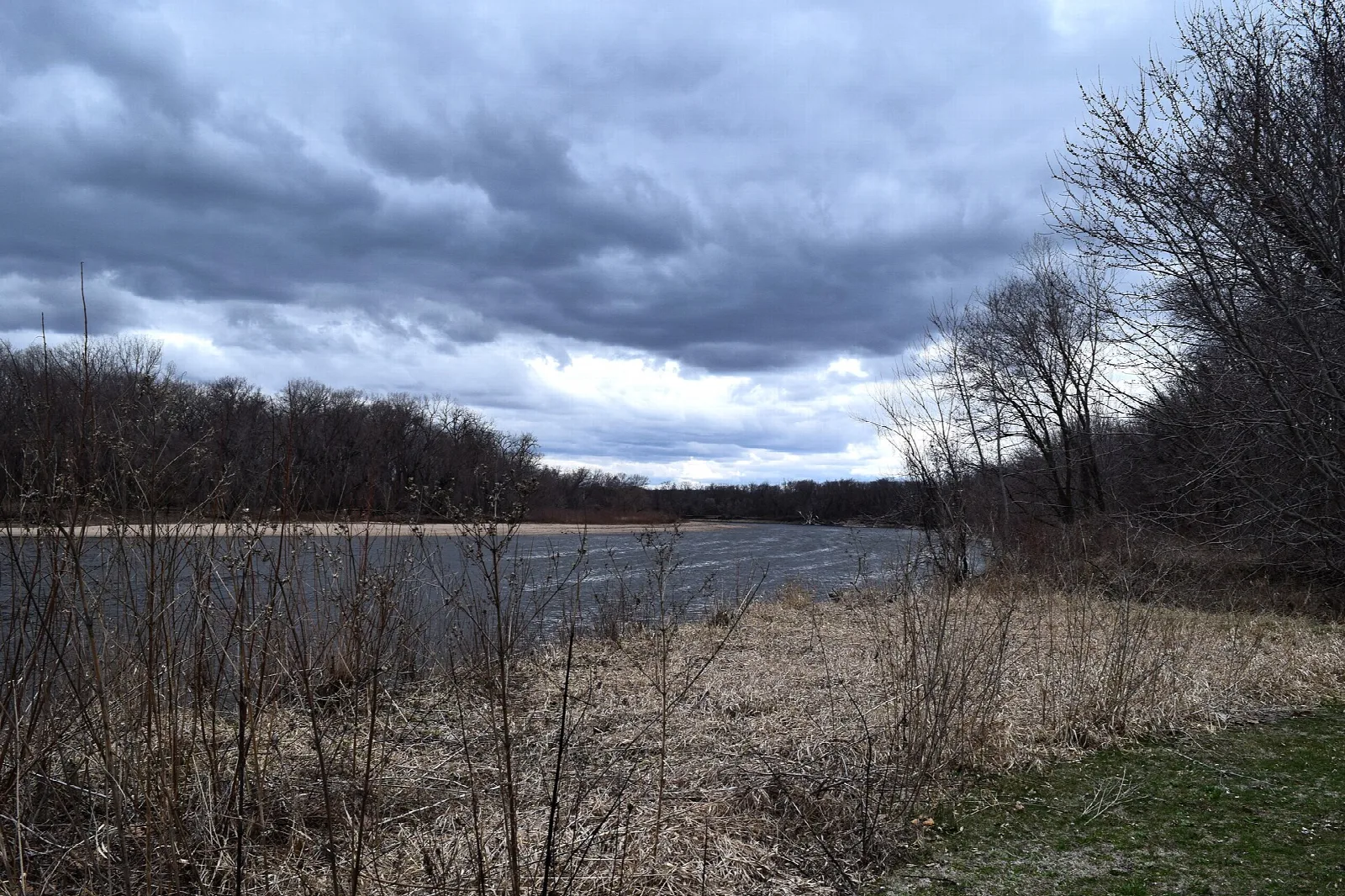 Wide river under a stormy sky with bare trees and dry grass along the bank
