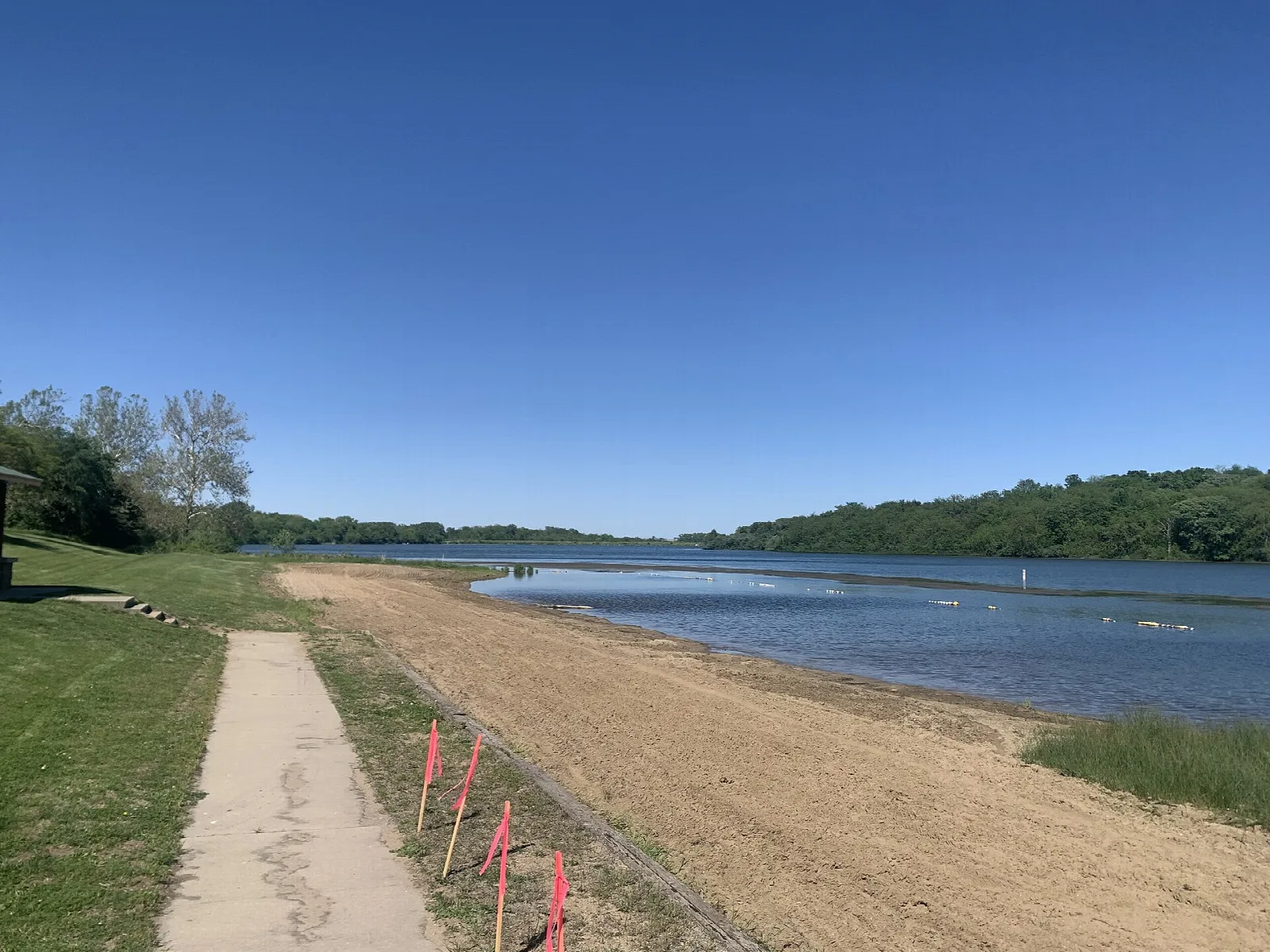 Sandy beach and a paved path beside a calm lake under a blue sky