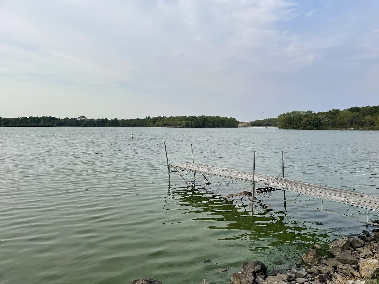 Lake under a hazy sky with a narrow dock reaching into the water and rocks at the shore