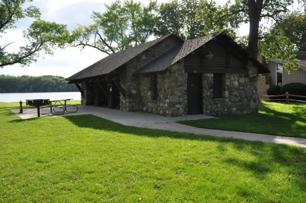 Stone picnic pavilion with a shingled roof on a green lawn beside the lake