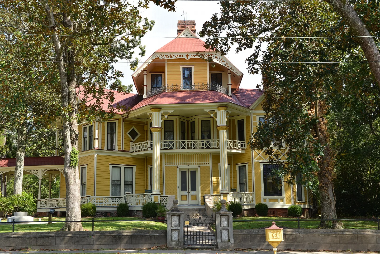 Yellow Victorian house with a tower and wraparound porches behind trees