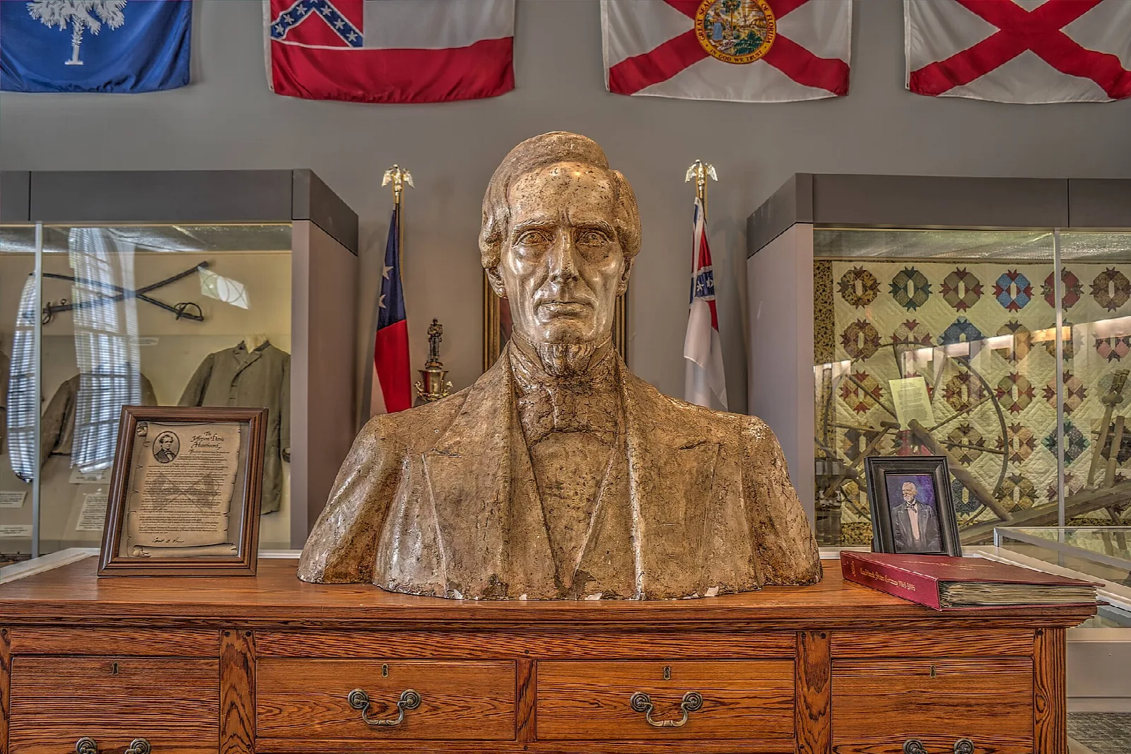 Bronze-colored bust on a wooden dresser, with flags and display cases behind it