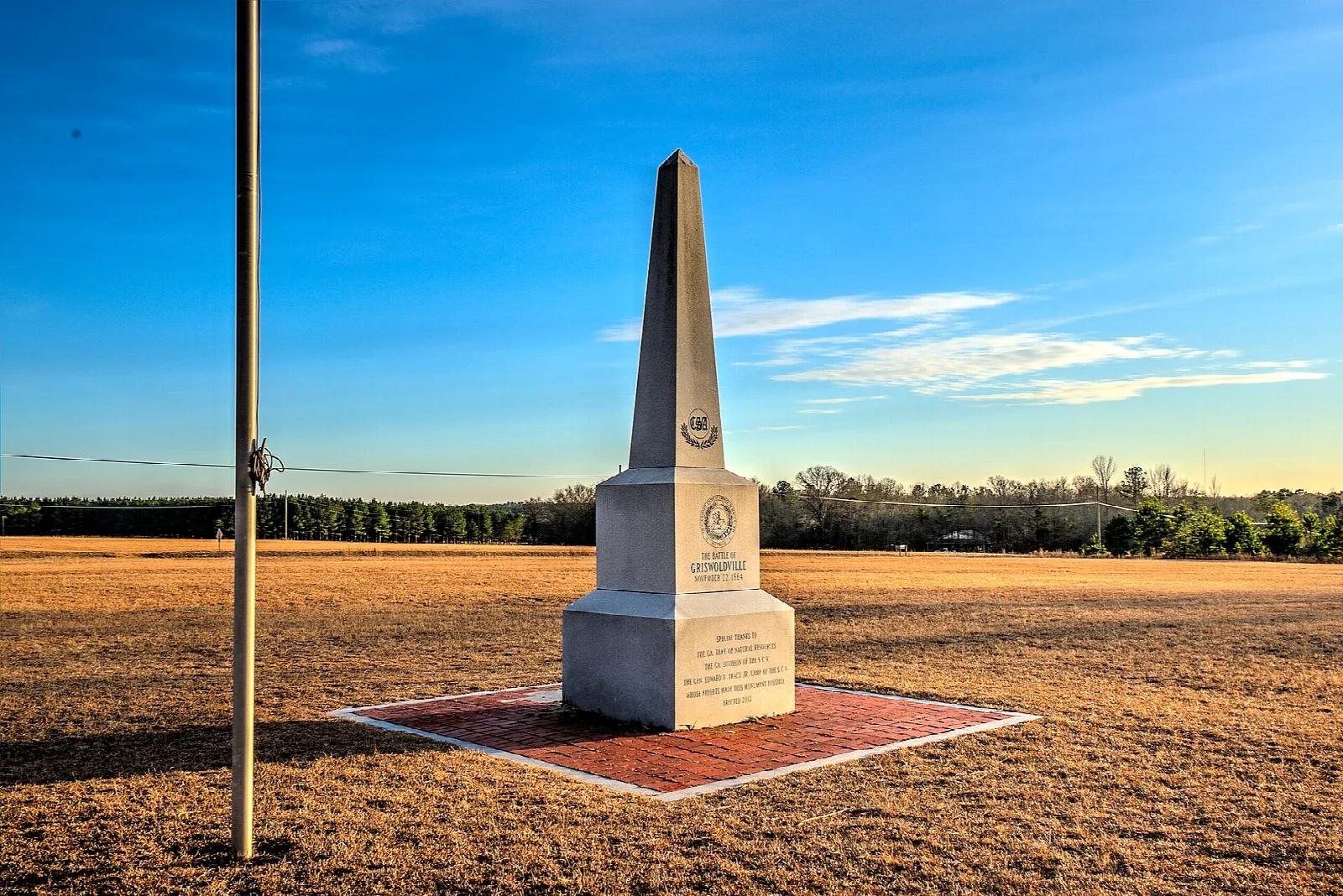 White stone obelisk on a brick pad in an open field of dry grass under a blue sky