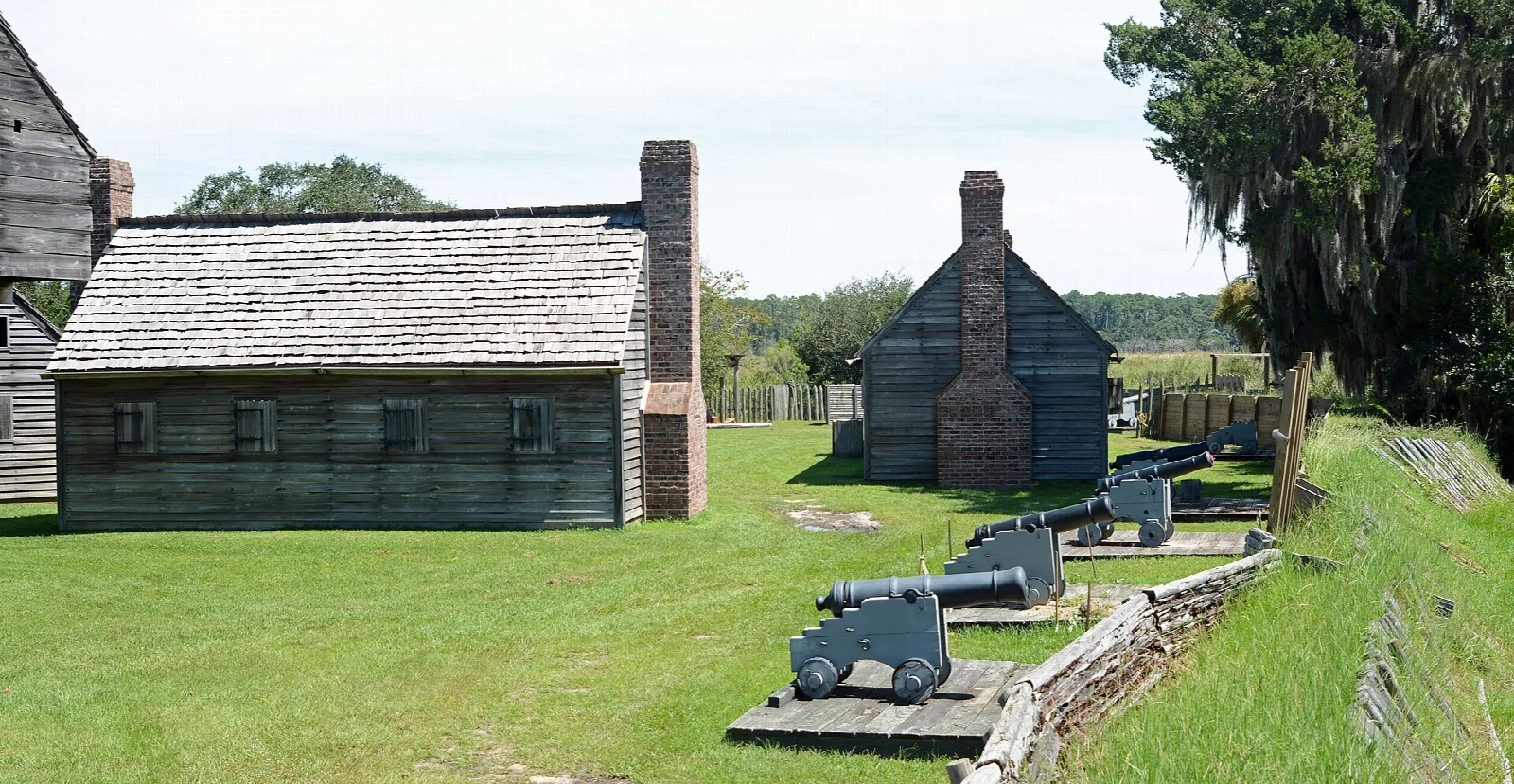 Wooden buildings with brick chimneys beside a grassy wall with cannons