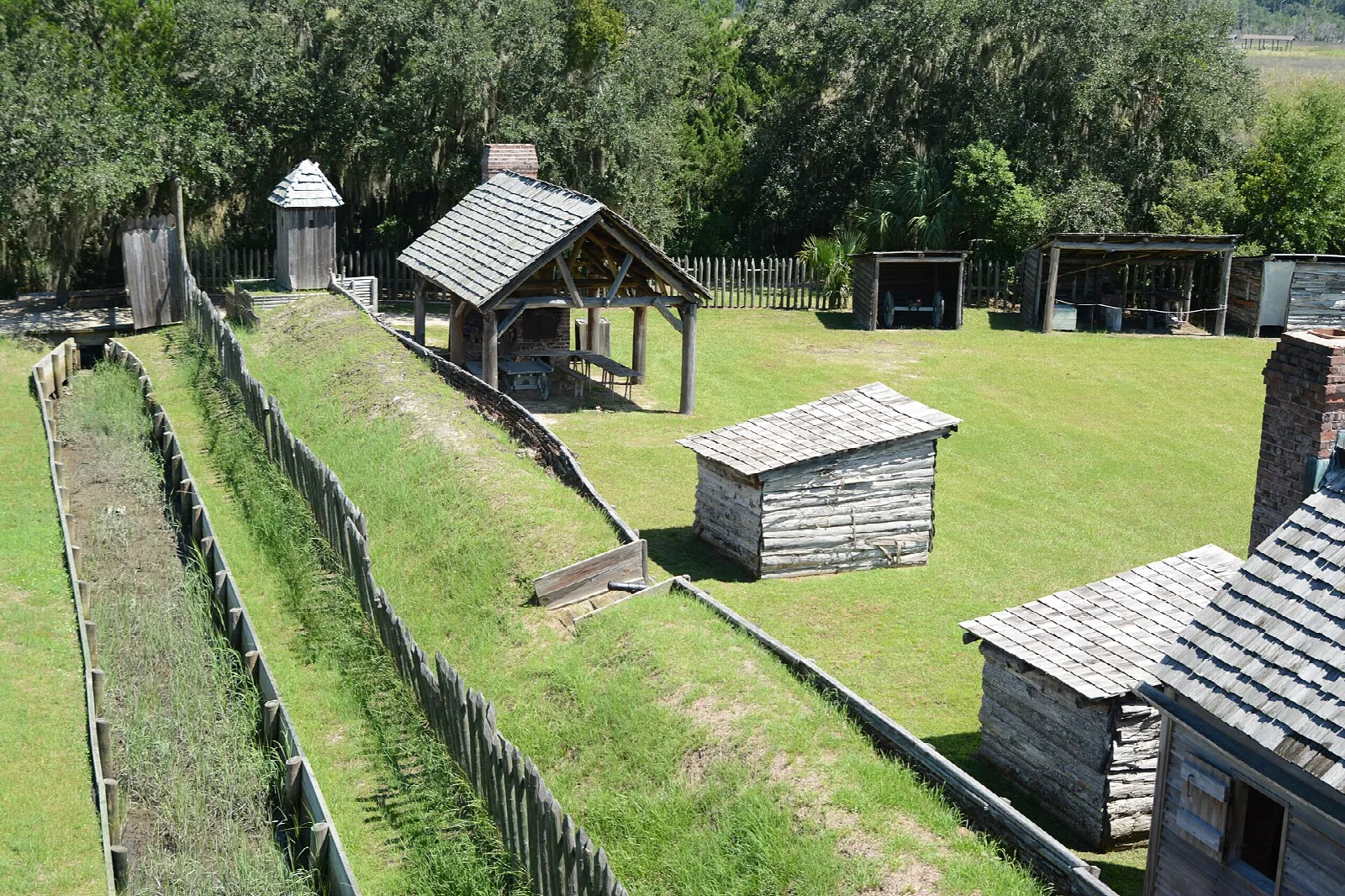 Grassy moat and earthen wall lined with log stakes, with small wooden buildings inside the fort