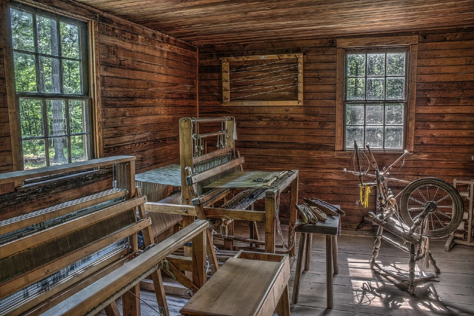Wood-paneled room with a loom and spinning wheel by two windows