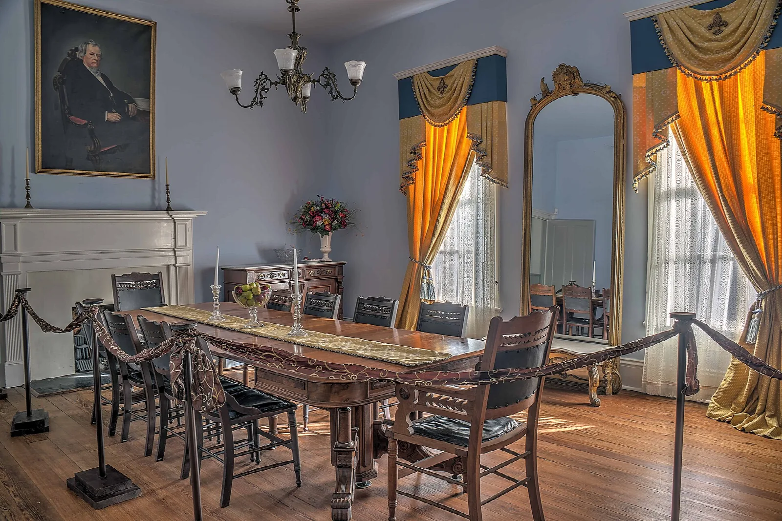 Dining room with a long wooden table, chairs, gold curtains and a portrait on the wall