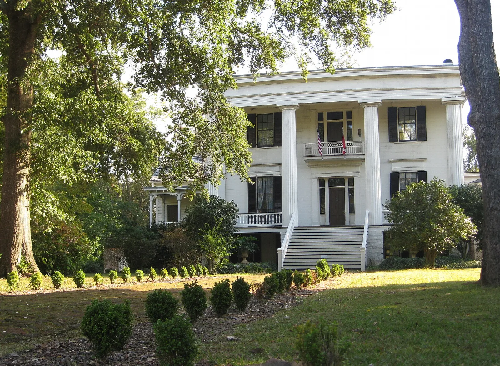 White columned house across a lawn with clipped shrubs under large trees