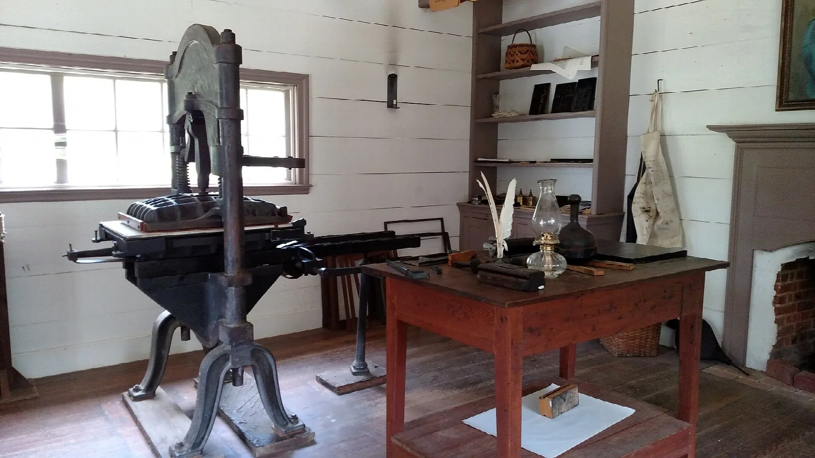 Old iron printing press beside a wooden desk in a bright white-walled room