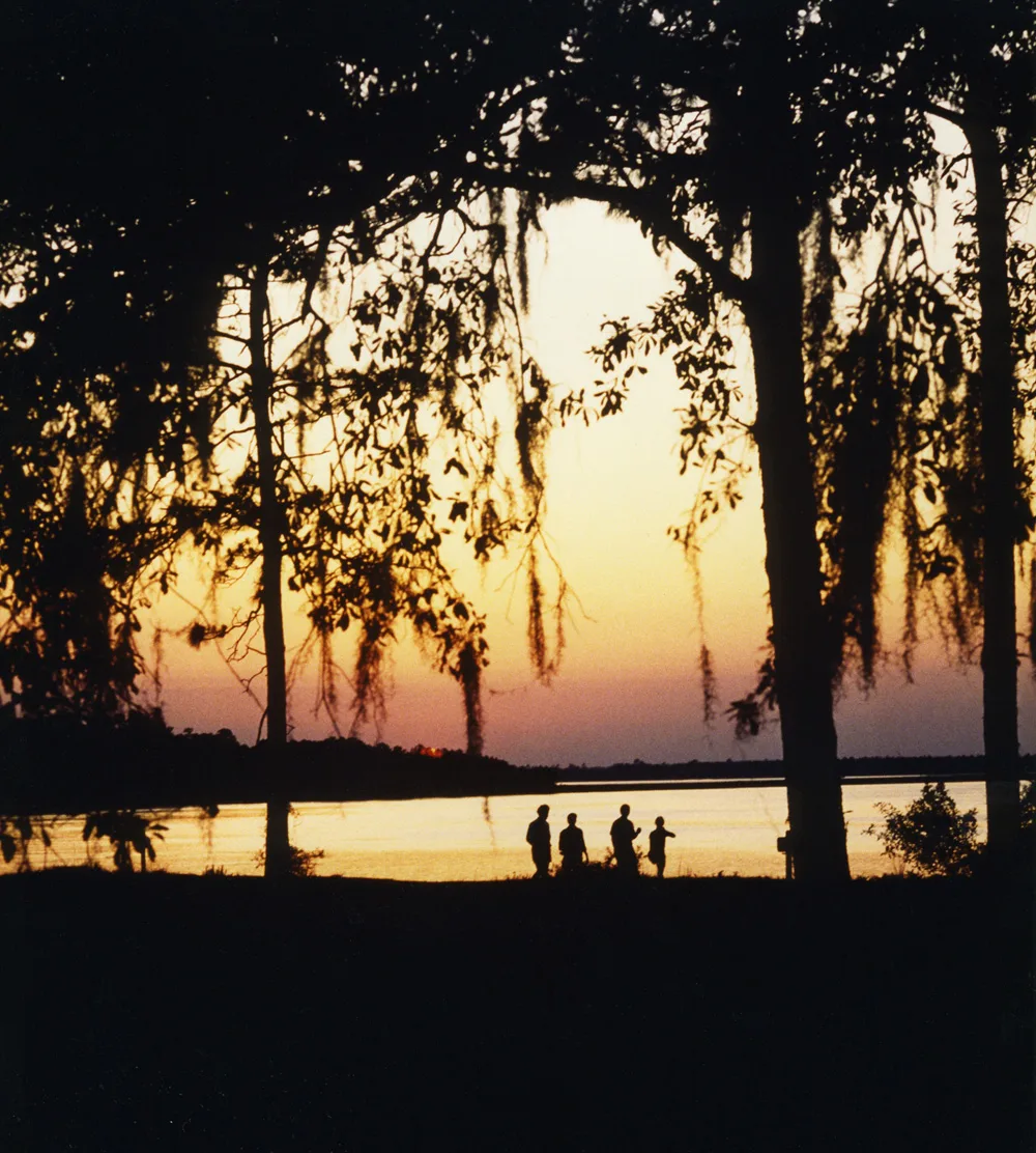 Sunset glowing over the river behind Spanish moss-draped trees, with a few people on the bank