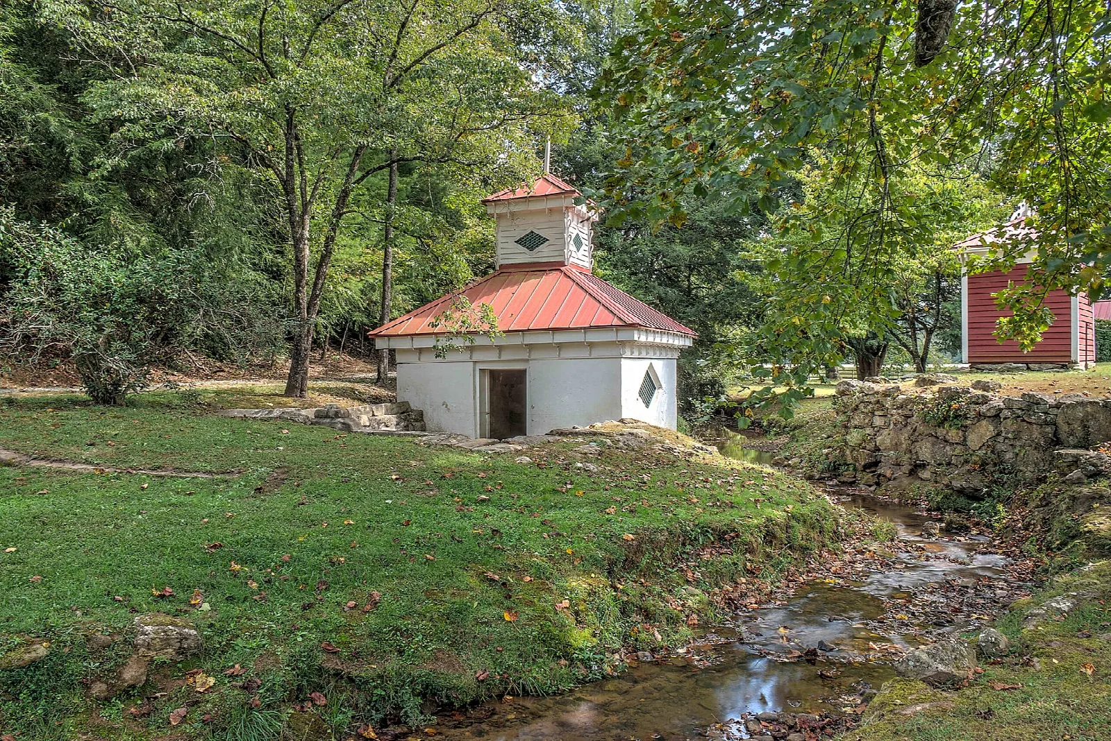 Small white spring house with a red roof beside a creek and lawn