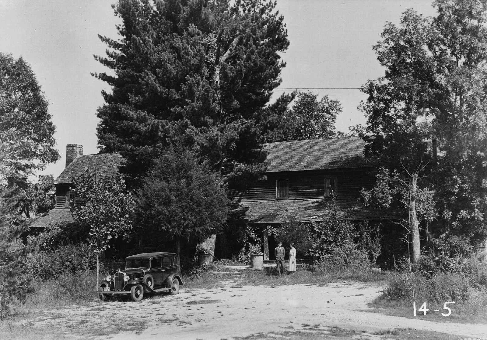 Black and white photo of the old inn beside tall trees with a vintage car in front