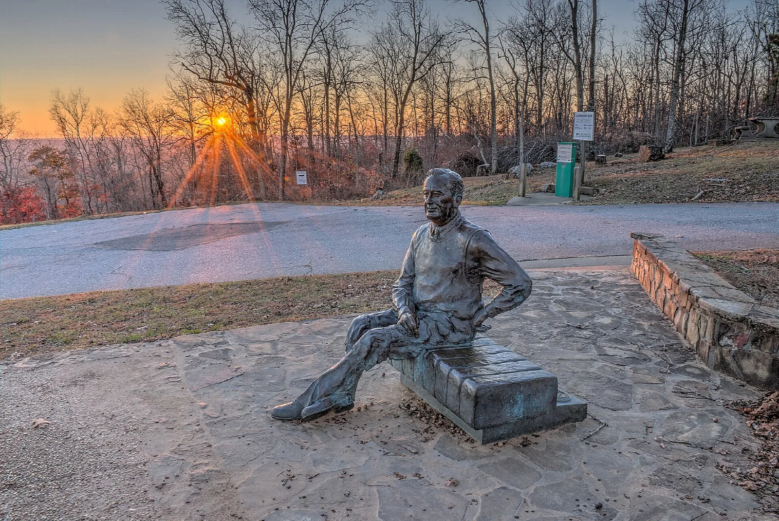 Bronze statue of a seated man at a stone overlook with the sun low on the horizon