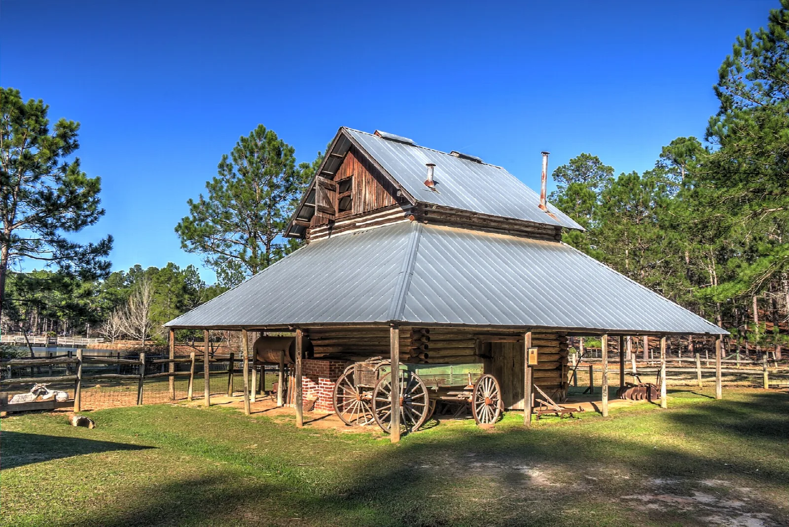 Weathered wooden tobacco barn with a metal roof and an old wagon under it, on a green lawn