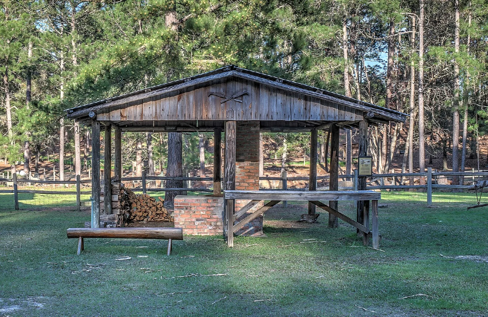 Open wooden shelter with a brick chimney and stacked firewood on a lawn