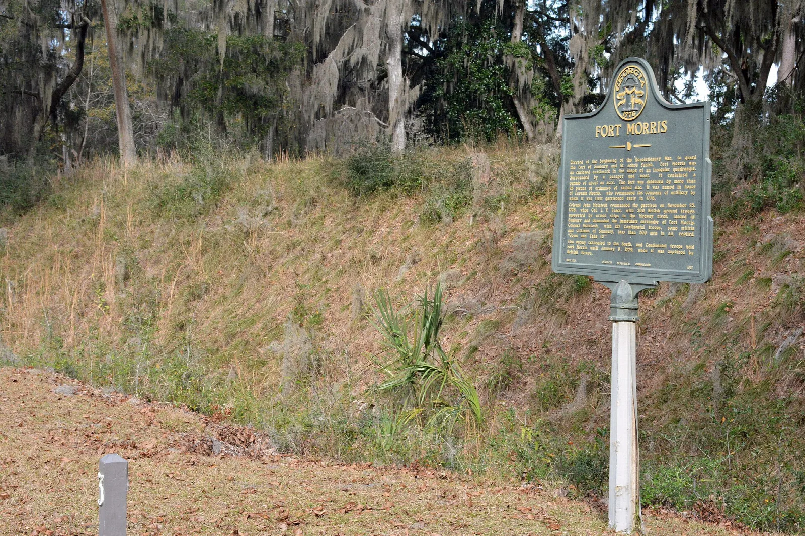 Grassy earthwork slope under moss-draped trees with a historical marker