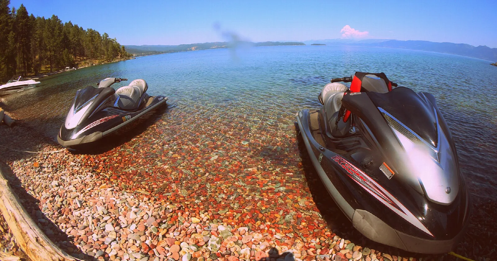 Two jet skis pulled up on a pebbly shore beside clear lake water