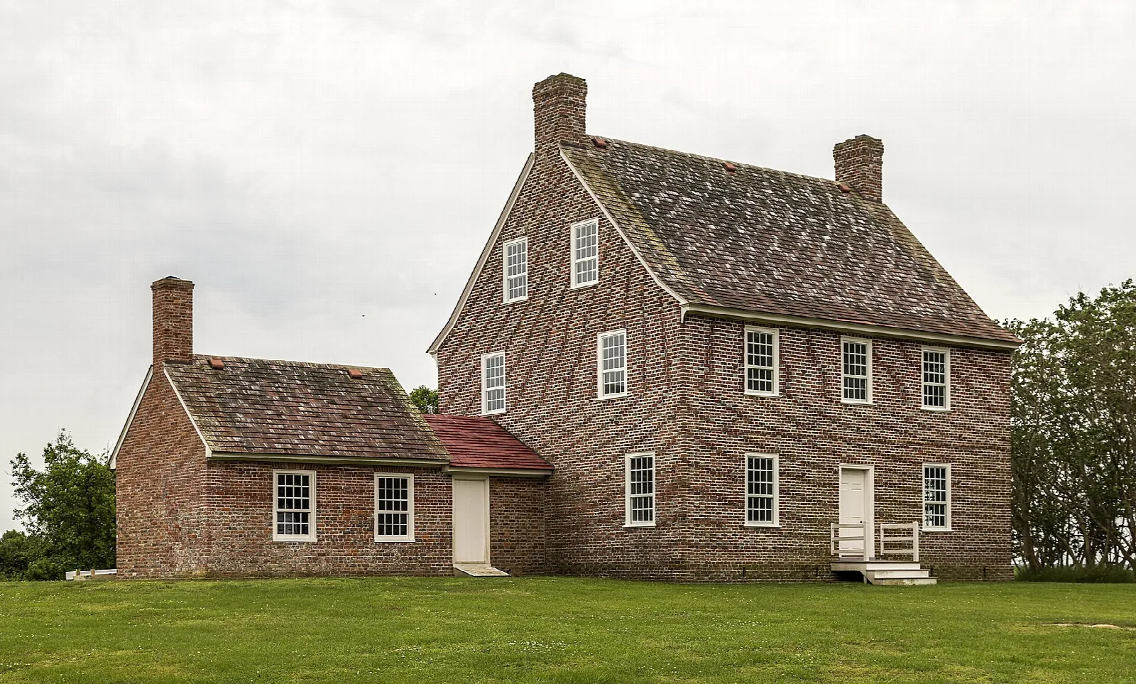 Two-story brick house with chimneys on a green lawn under a cloudy sky