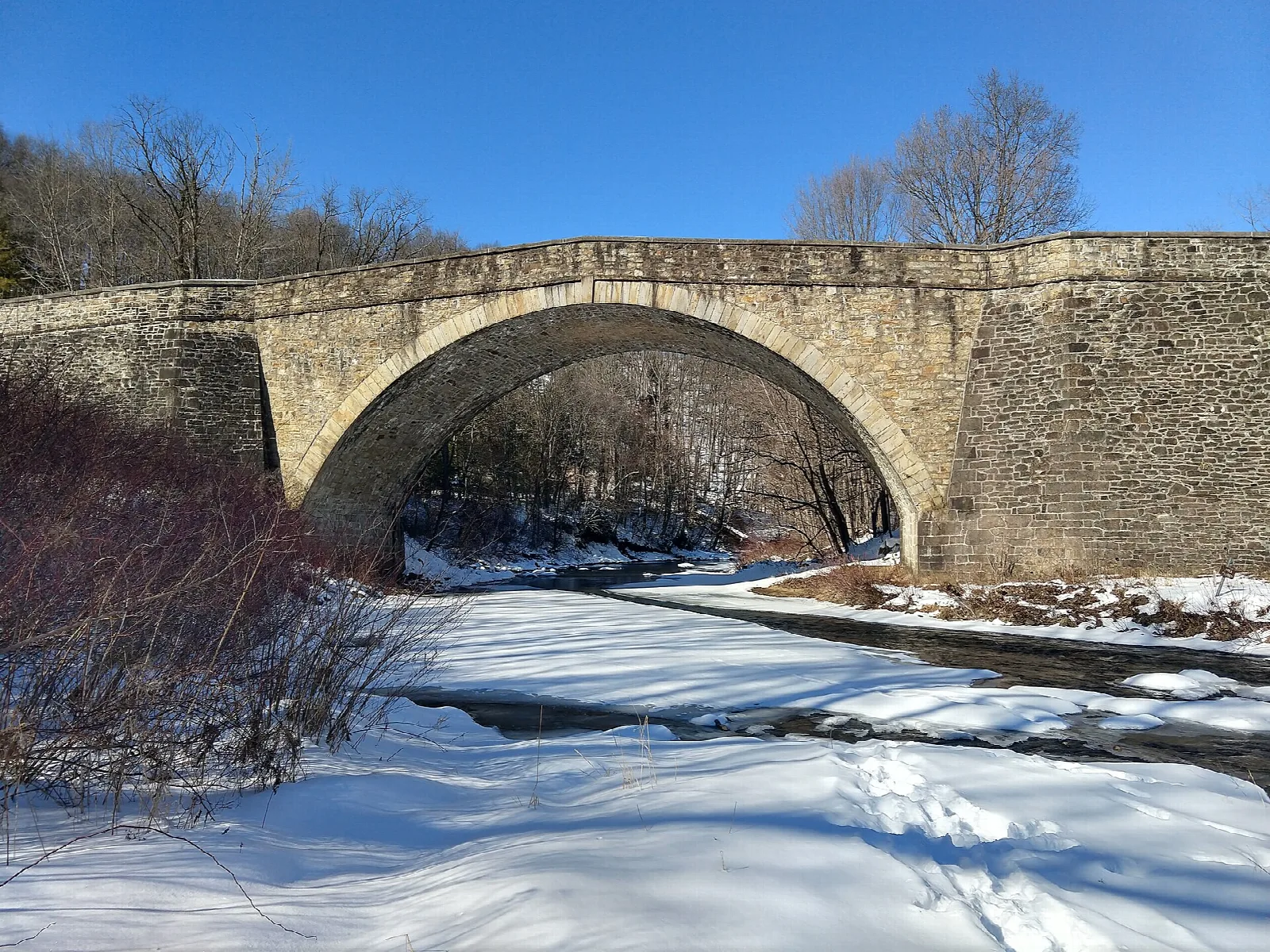 Stone arch bridge over a river with snow on the banks and blue sky
