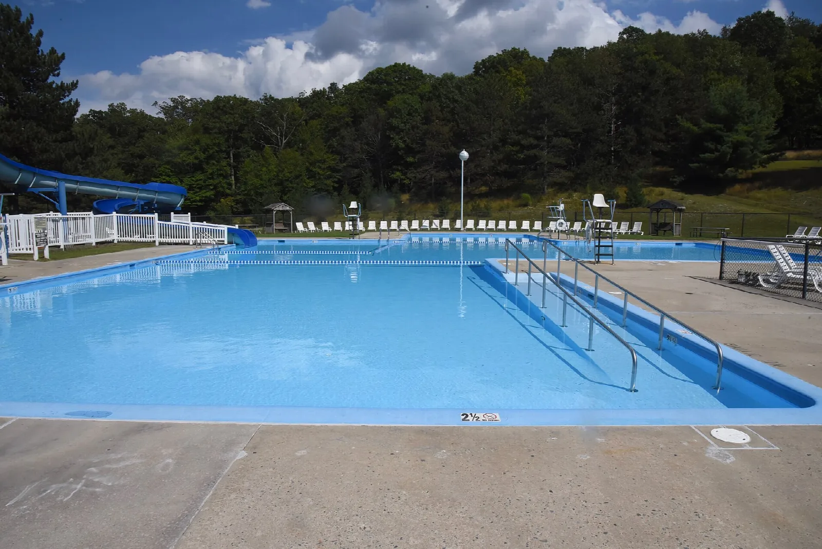 Large blue outdoor swimming pool with a water slide and wooded hills behind