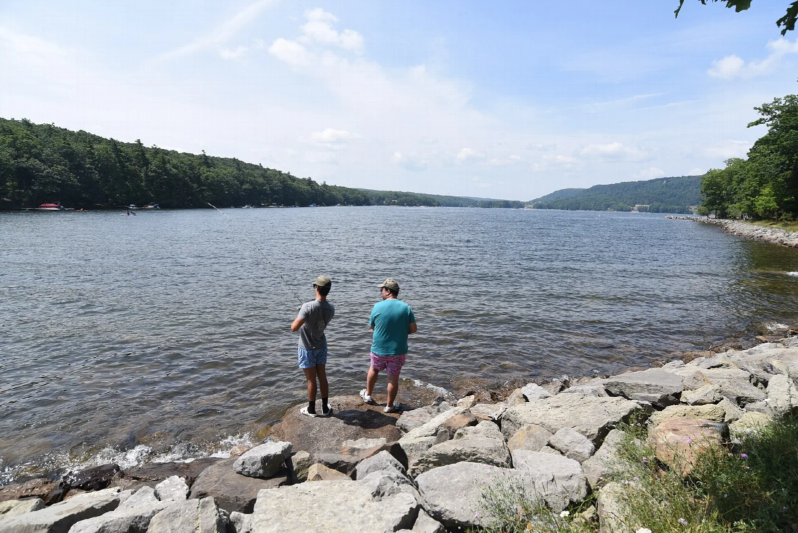 Two people standing on a rocky lake shore with forested hills beyond