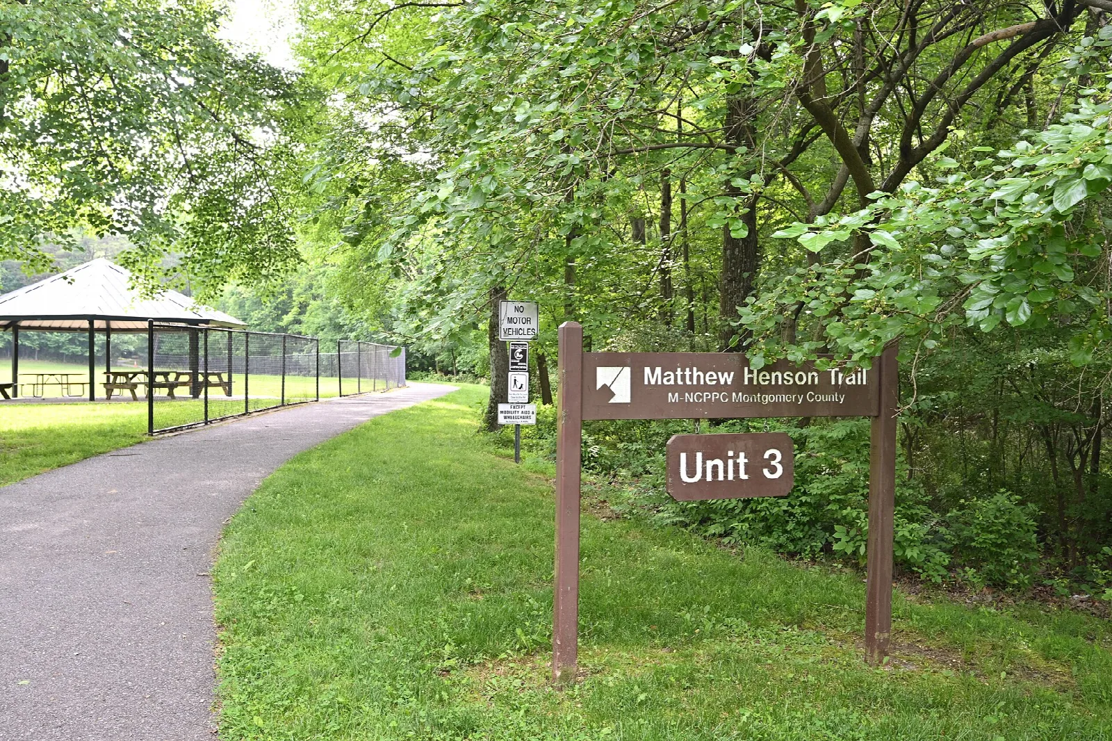 Paved trail beside a lawn and picnic shelter, with a Matthew Henson Trail Unit 3 sign