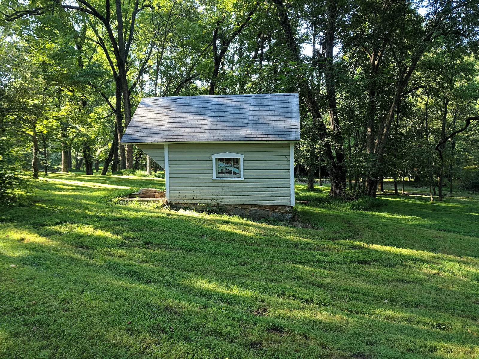 Small pale green outbuilding on a grassy lawn in the woods