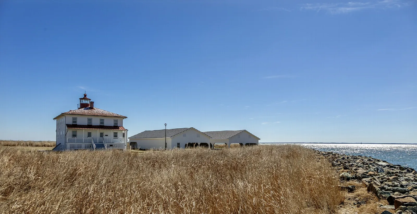 White lighthouse keeper's house with red roof behind tall dry grass and rocks