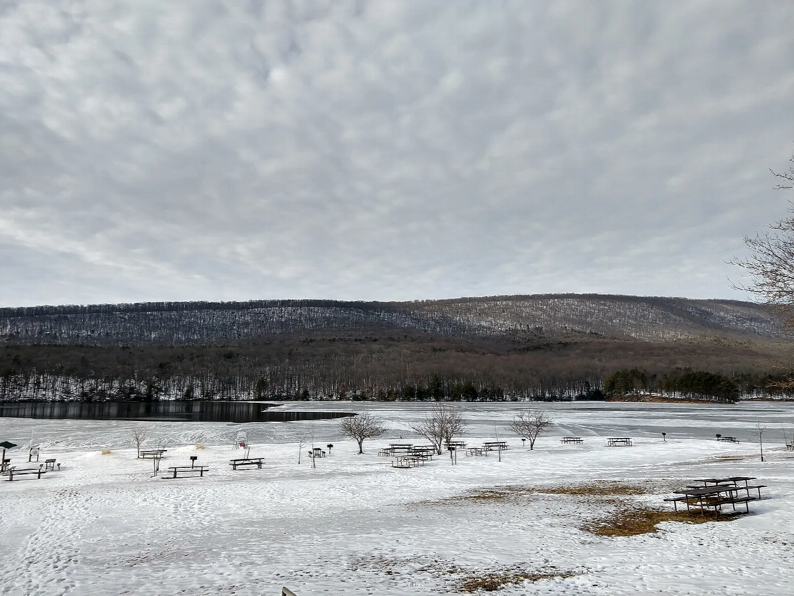 Snow-covered beach and picnic tables beside a lake with hills behind