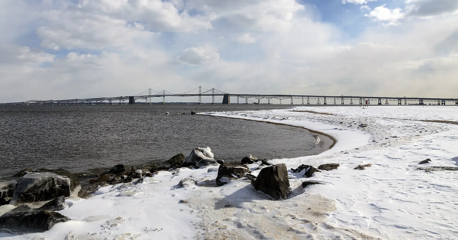 Snowy beach and rocks beside gray water with the long Bay Bridge in the distance