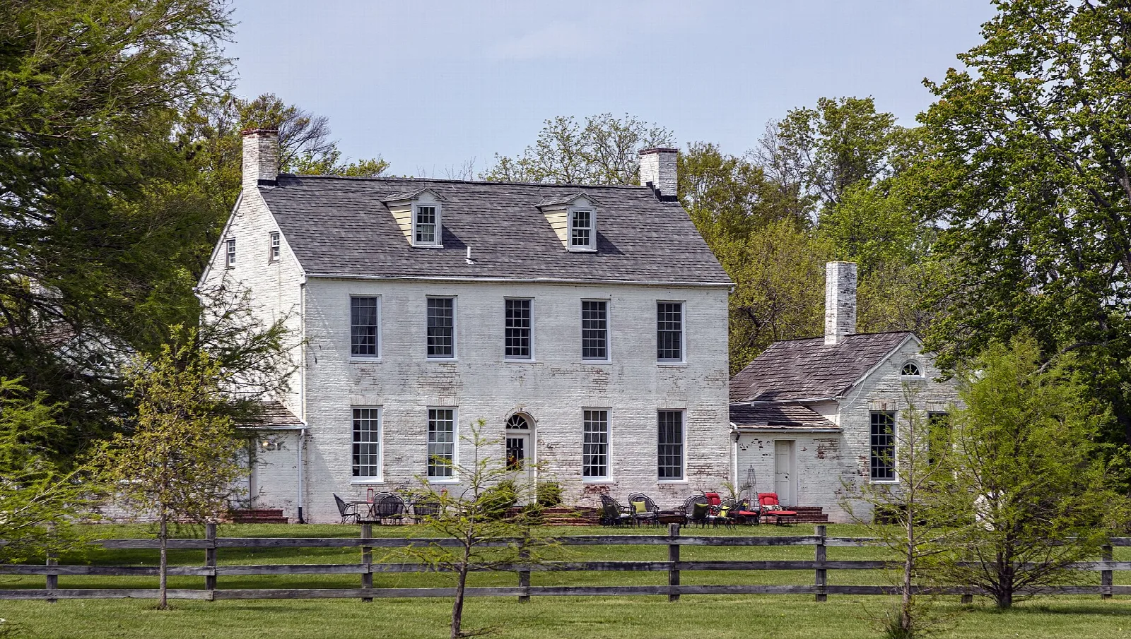White farmhouse with chimneys across a grassy field behind a split-rail fence