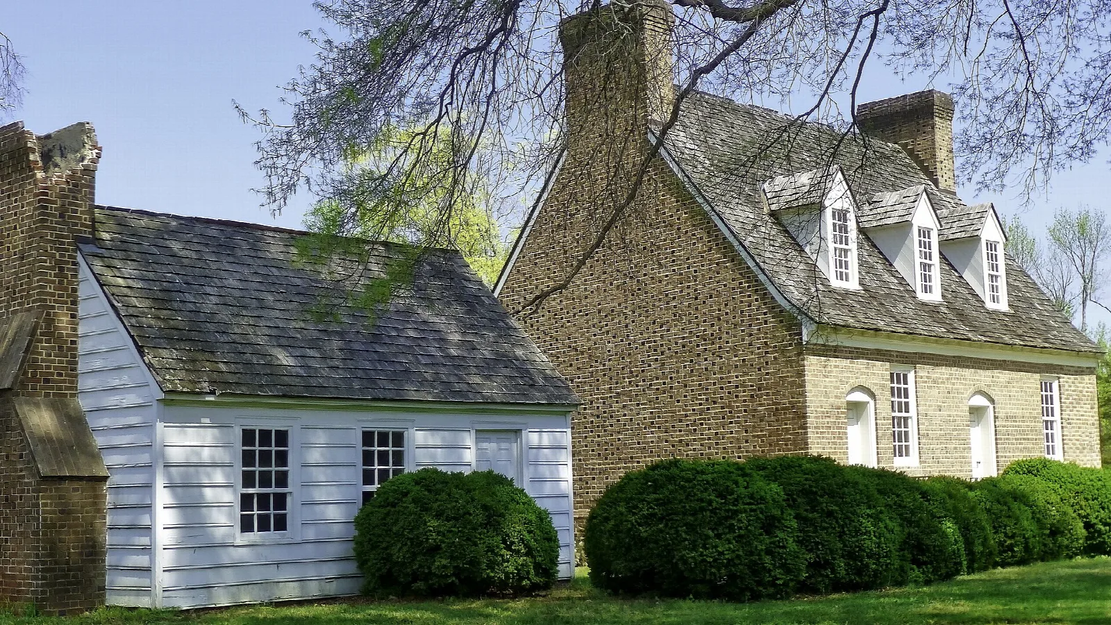 Brick house with dormers and a white wooden wing behind shrubs