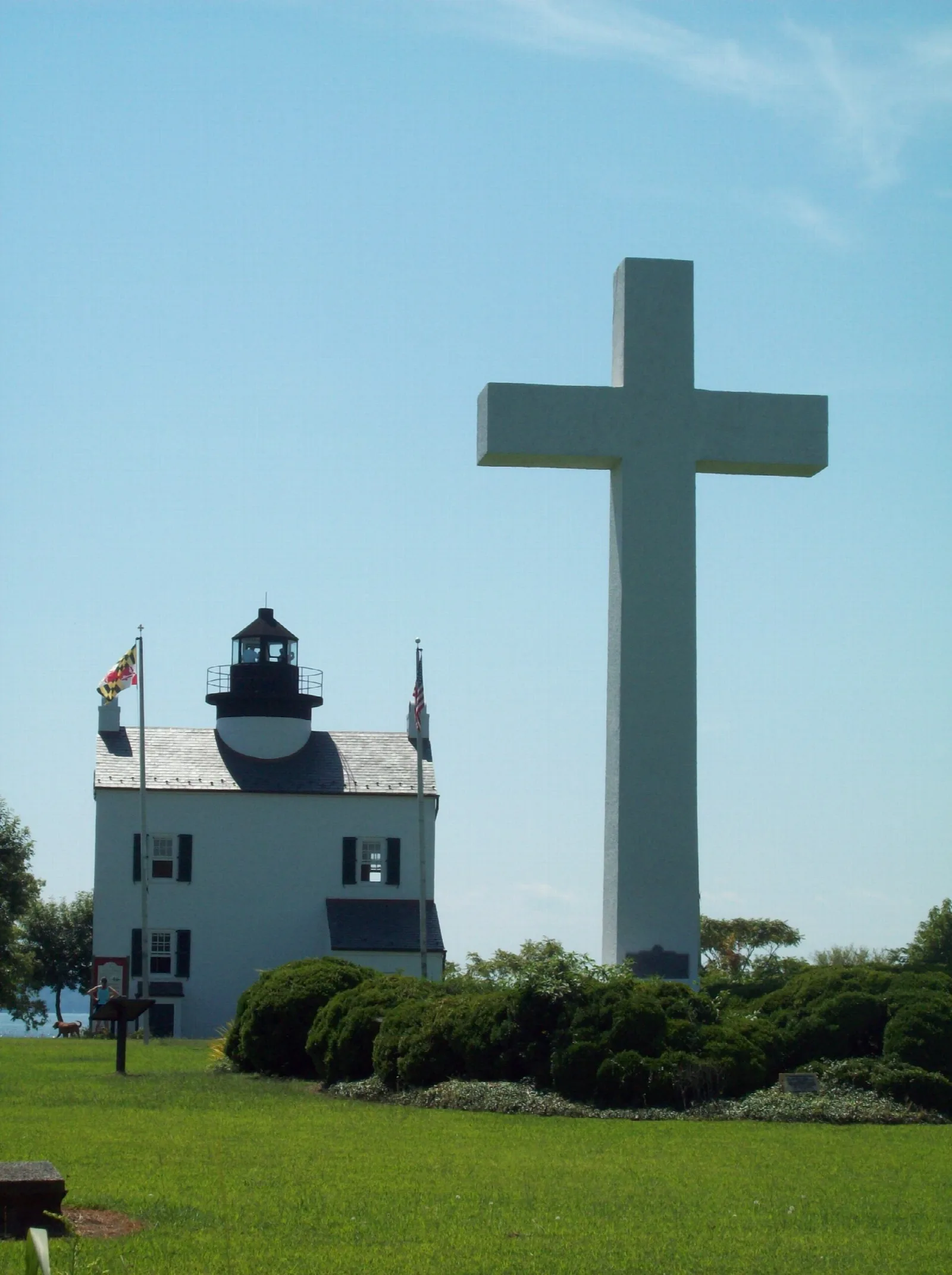 Tall white cross beside a white lighthouse building on a lawn