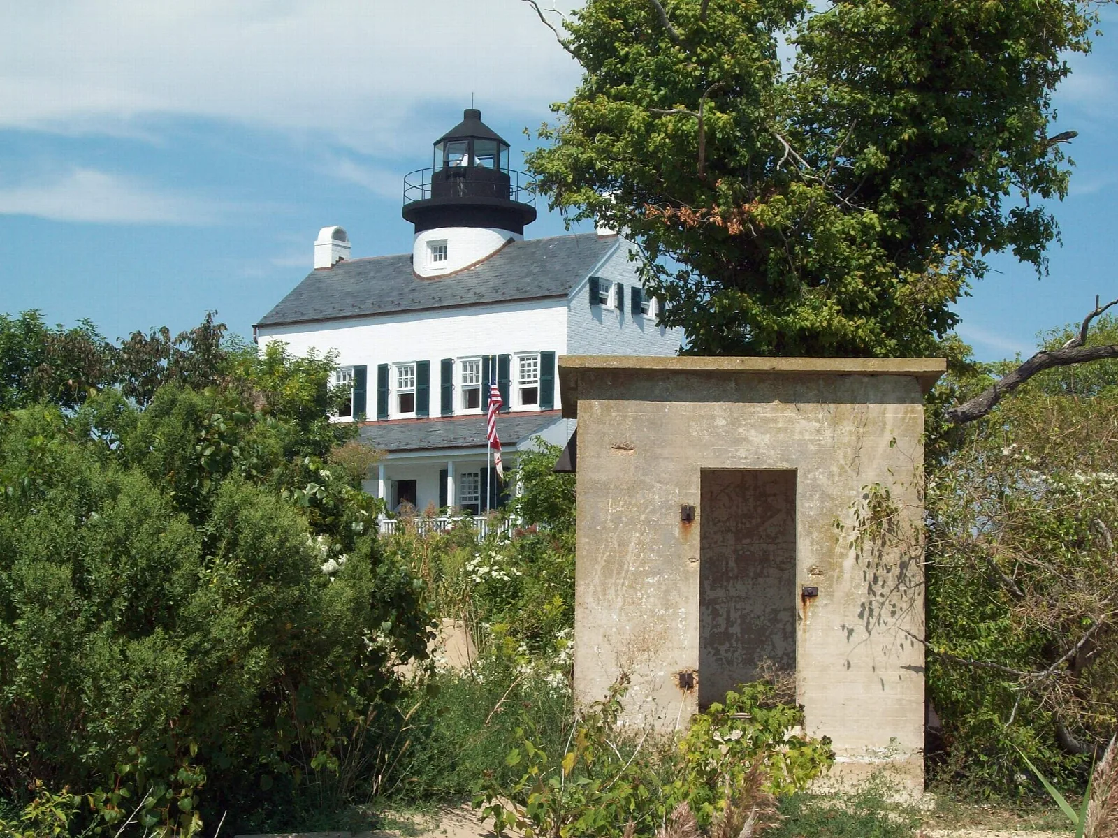 Lighthouse behind a stone shed and shrubs under blue sky