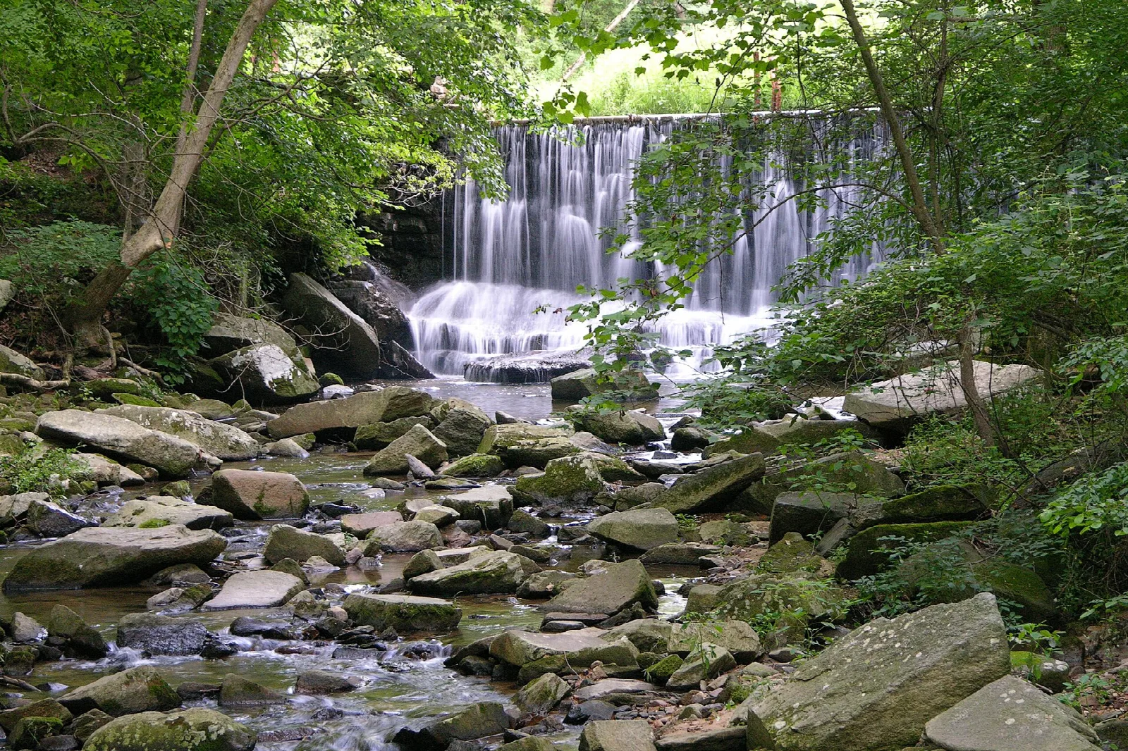 Waterfall dropping over a low dam into a rocky, wooded stream