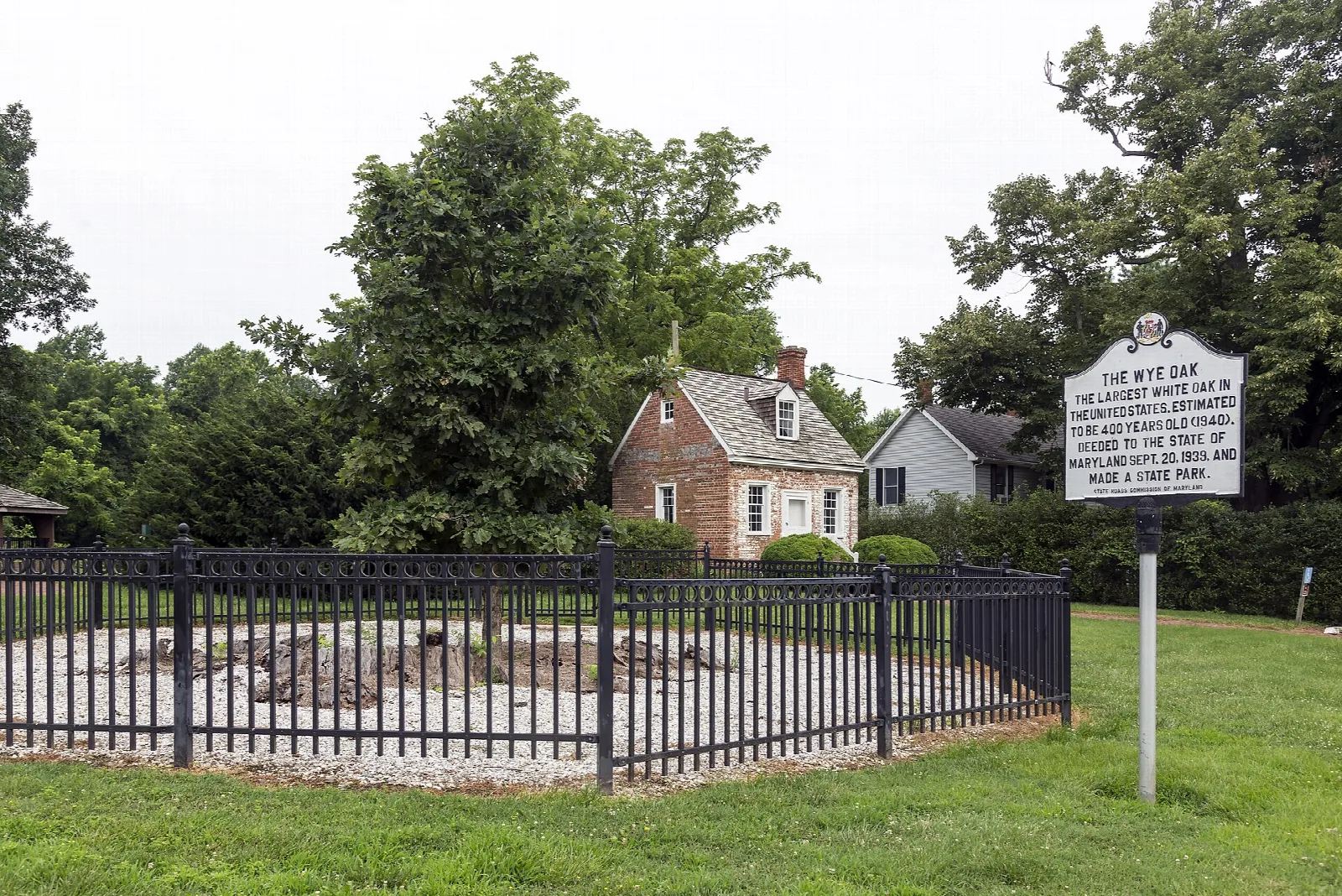 Fenced circle at the original tree site beside a brick schoolhouse and a marker sign