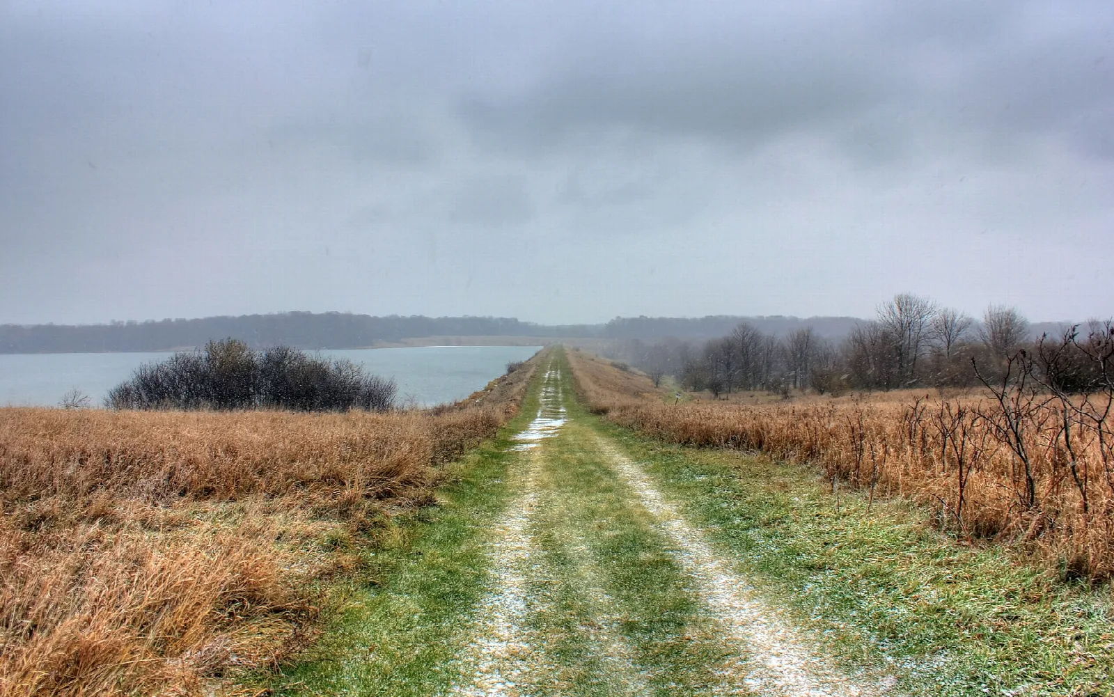 Grassy trail running through dry prairie toward a lake under gray sky