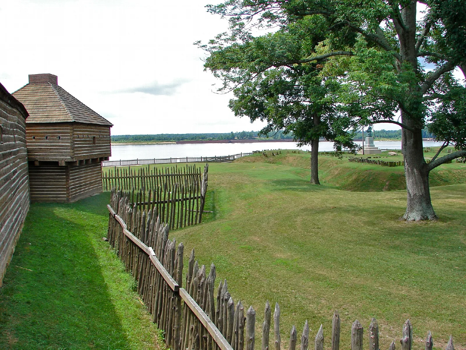 Grassy slope and wooden palisade fence beside a reconstructed log blockhouse, with a wide river beyond