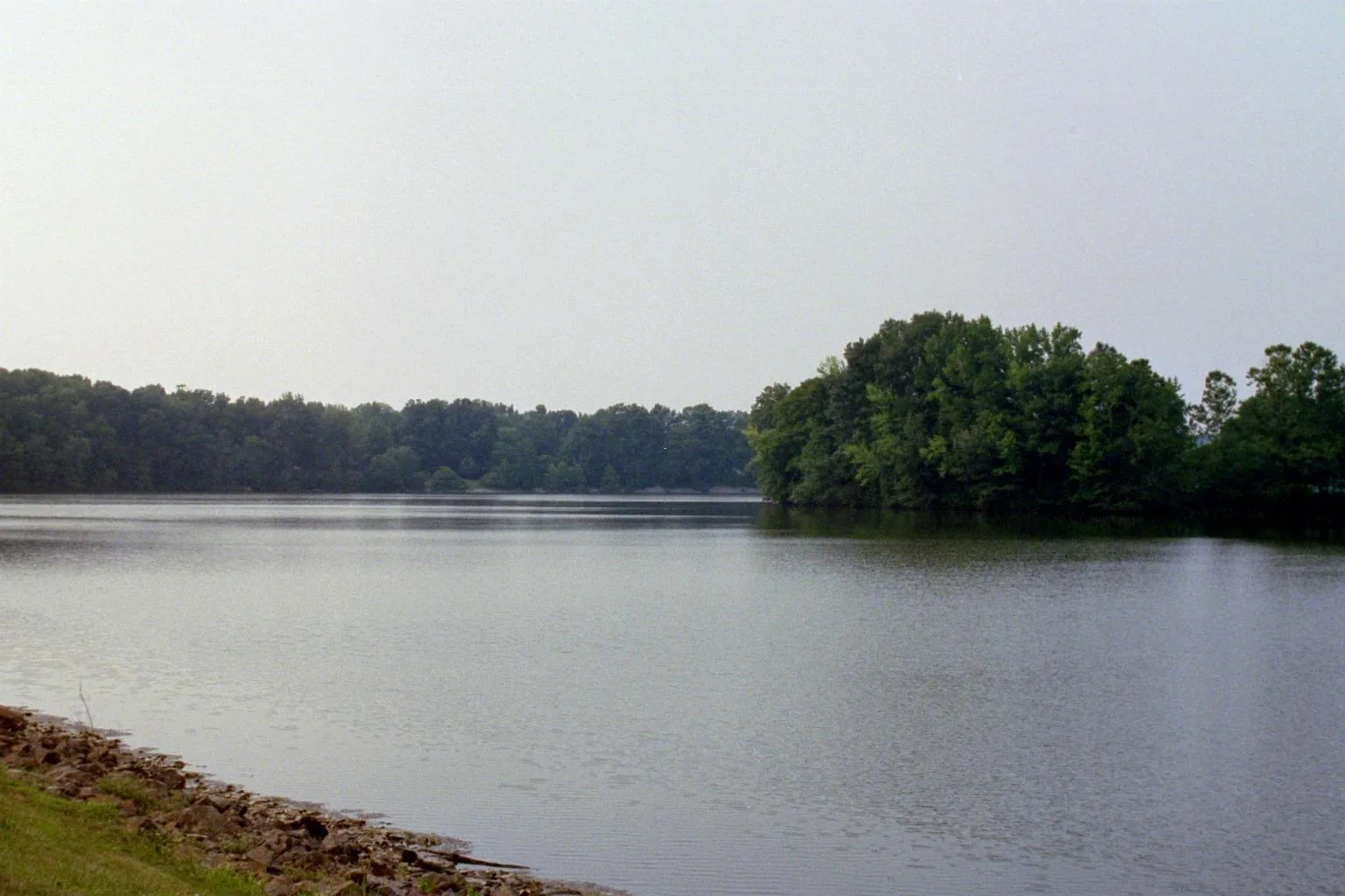 Calm lake with a tree-covered point and a rocky shoreline under a hazy sky