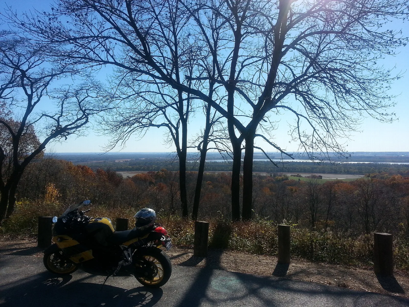 Overlook through bare trees toward a distant river valley, with a parked motorcycle