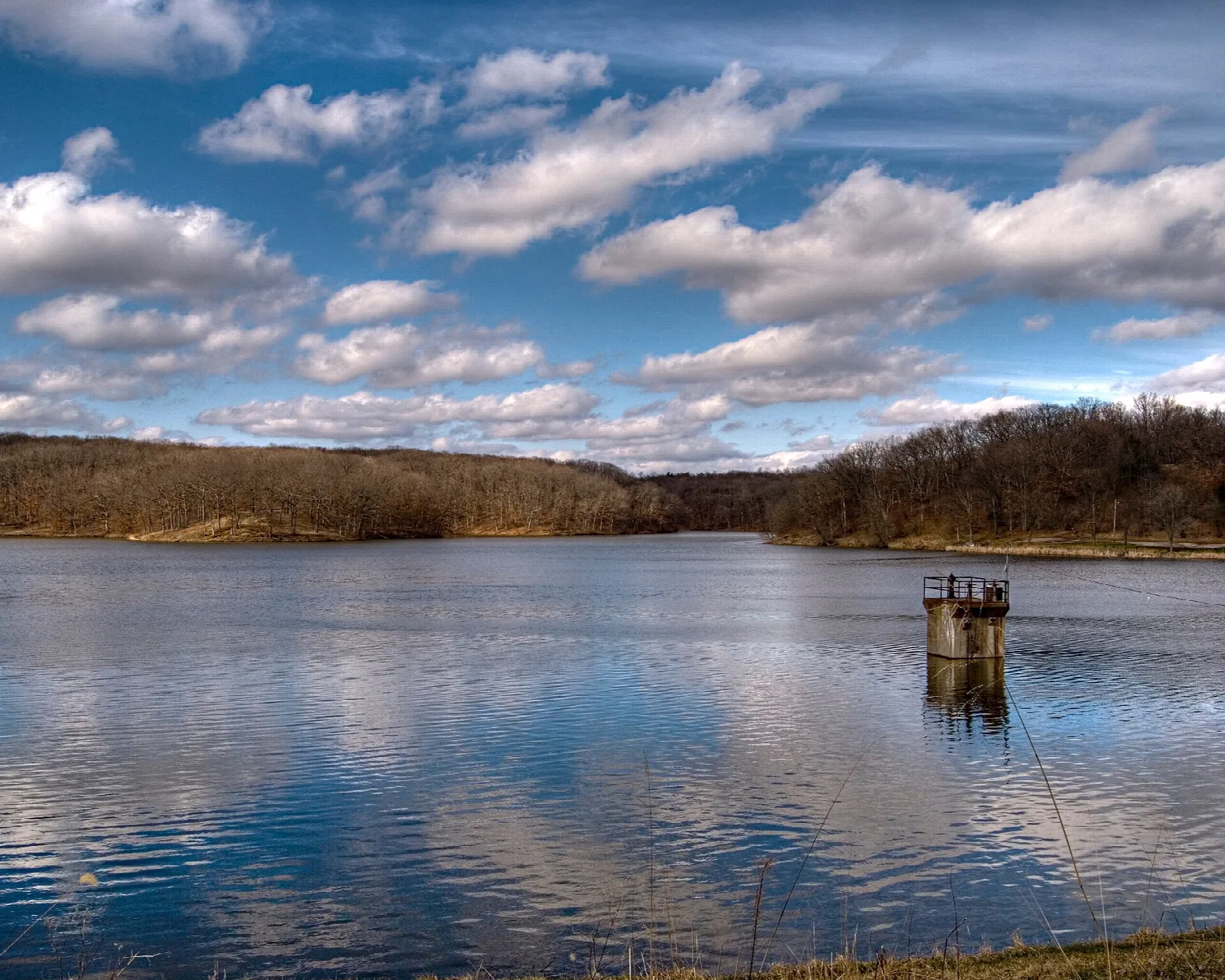 Wide lake with reflected clouds, wooded far shore, and a small concrete tower in the water