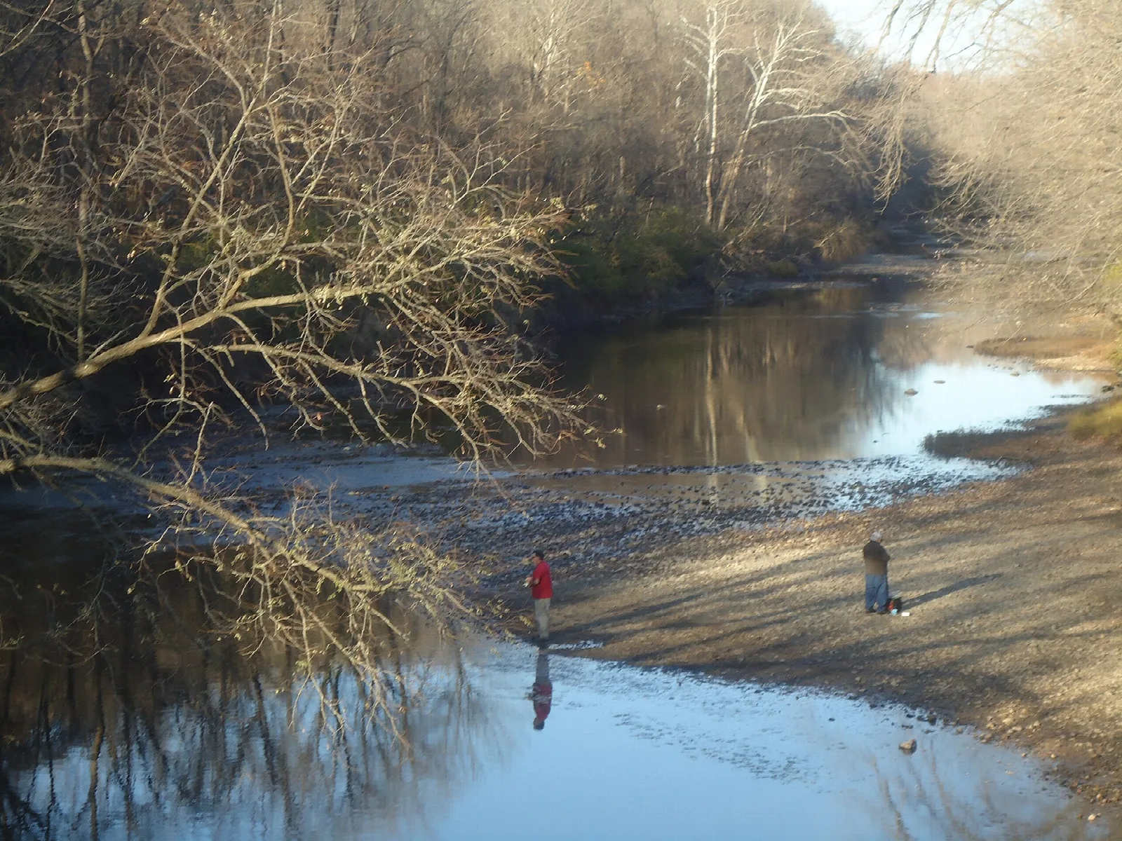 Two people fishing along a quiet river bend lined with bare trees