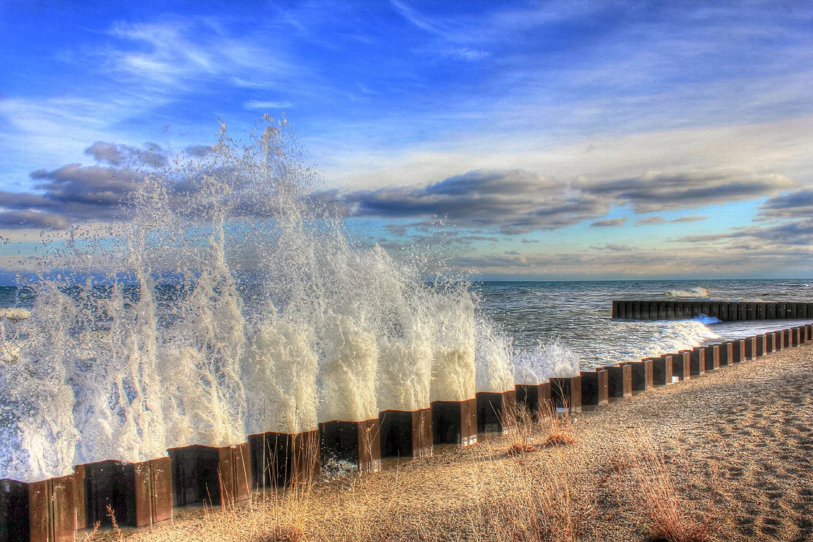 Waves splashing over a row of wooden posts on a sandy lake shore