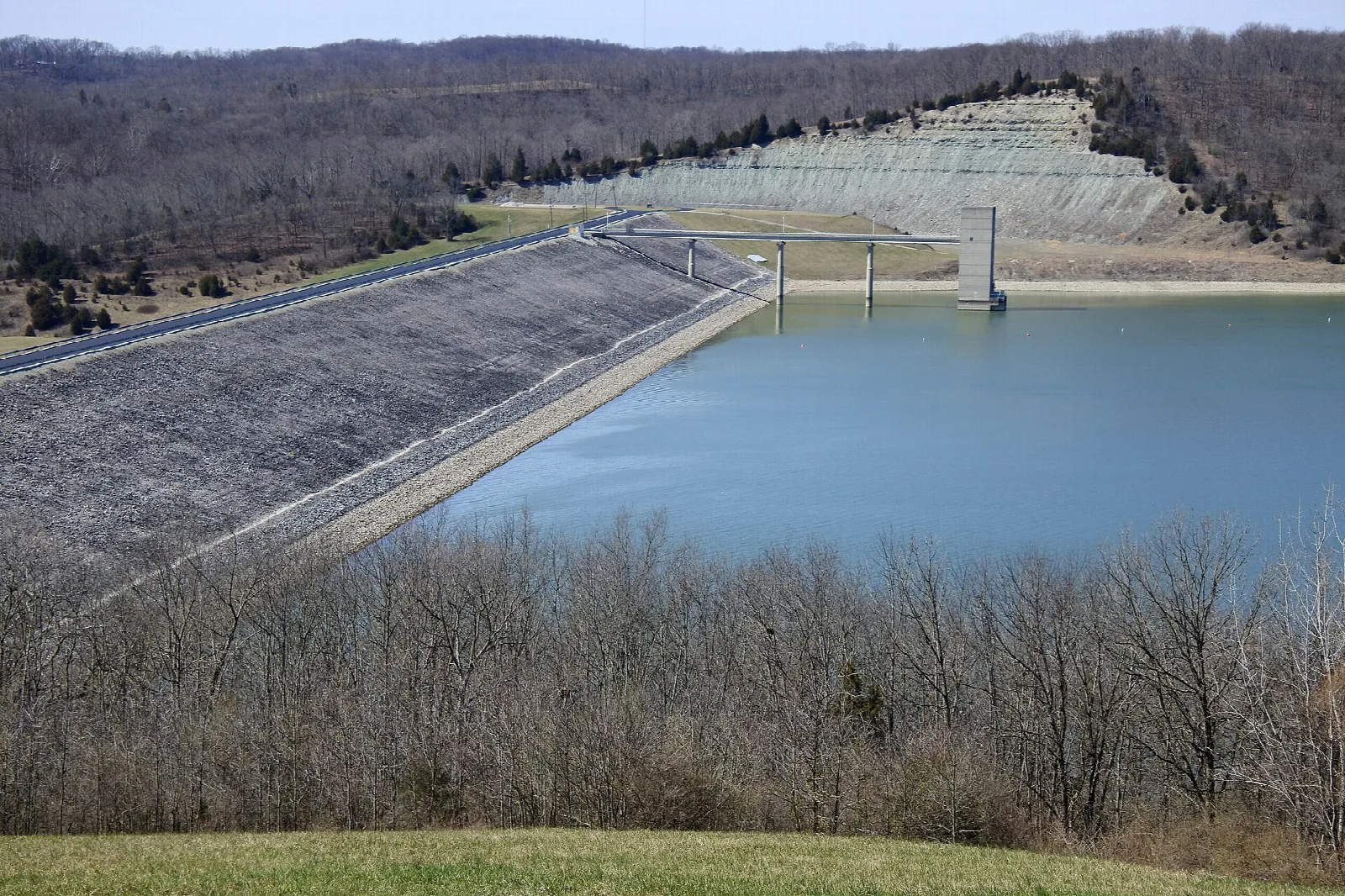 Earth dam beside a blue reservoir, seen from a grassy overlook with bare trees