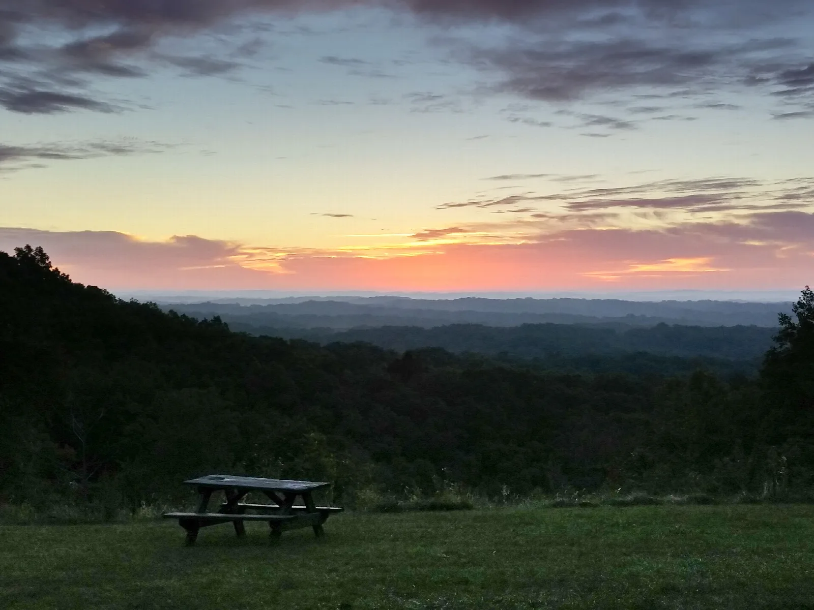 Picnic table on a grassy overlook at sunrise above dark ridges