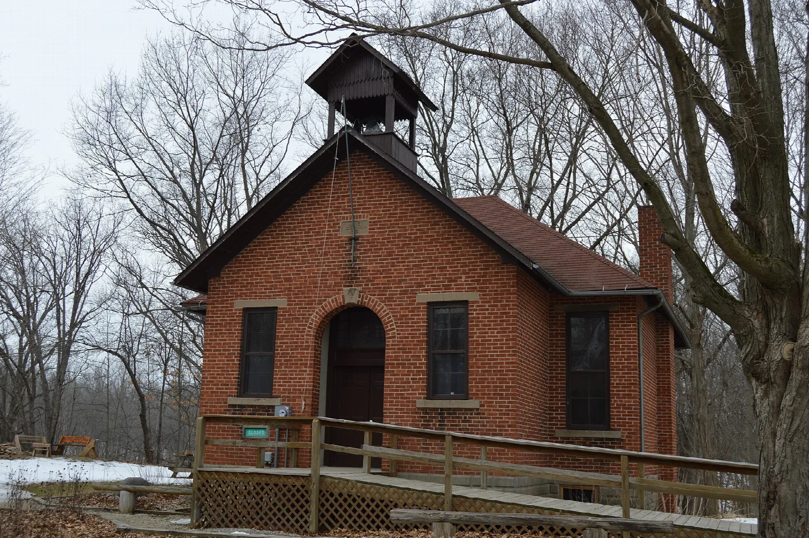 Red brick one-room schoolhouse with a bell cupola and a wooden ramp, among bare trees