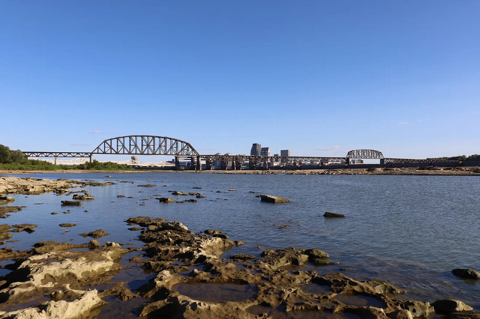 Railroad bridge over the river with the city skyline beyond, from a rocky shore