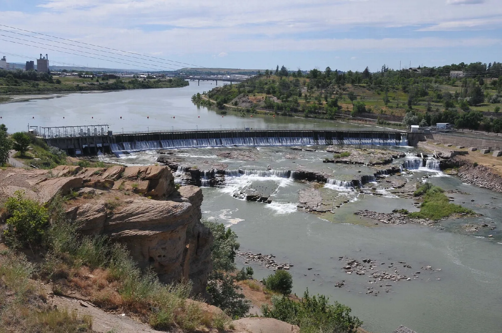 Wide river with a low weir and rapids below rocky bluffs, industrial buildings far off