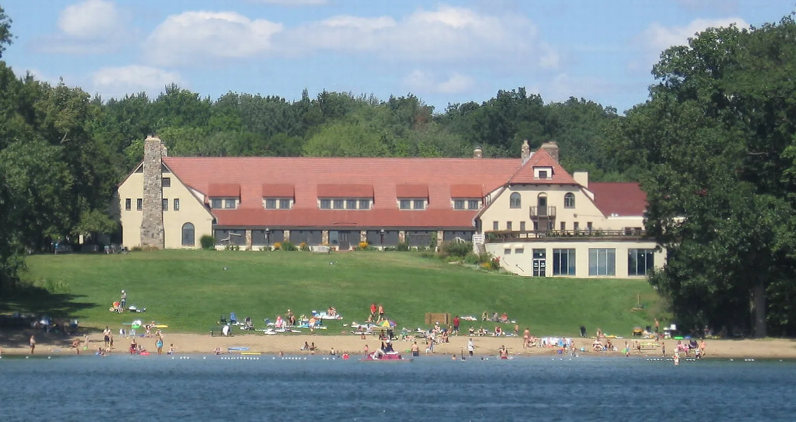 Large red-roofed lodge on a grassy shore above a lake, with people on the beach