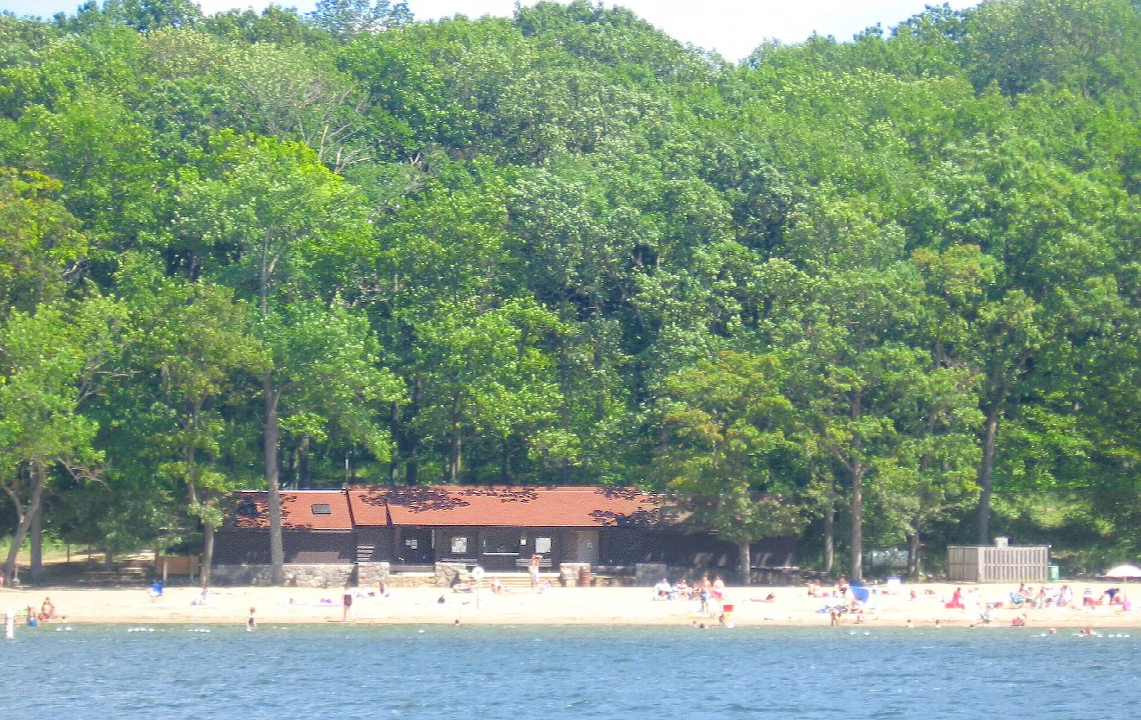 Low red-roofed beach house behind a sandy beach and swimmers, with wooded hills behind